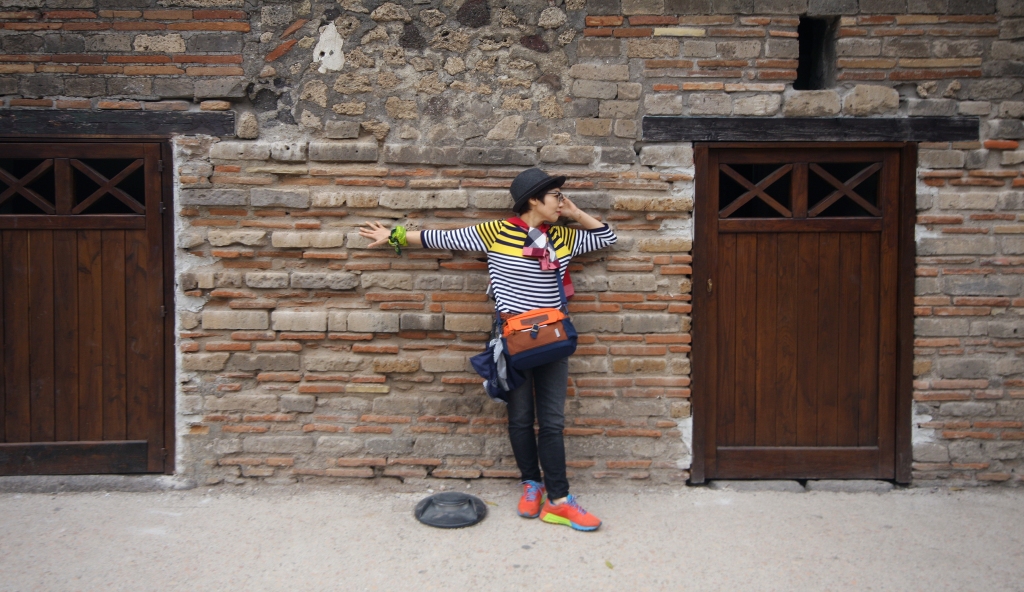 Photo is of a person in a striped, multicolored shirt, hat, pants, multicolored shoes, and bag around the shoulder. The person is posing in front of a stone wall with doors on both sides. The person is posing with the right arm stretched out straight along the wall and the left arm bent so that the hand is touching the face.