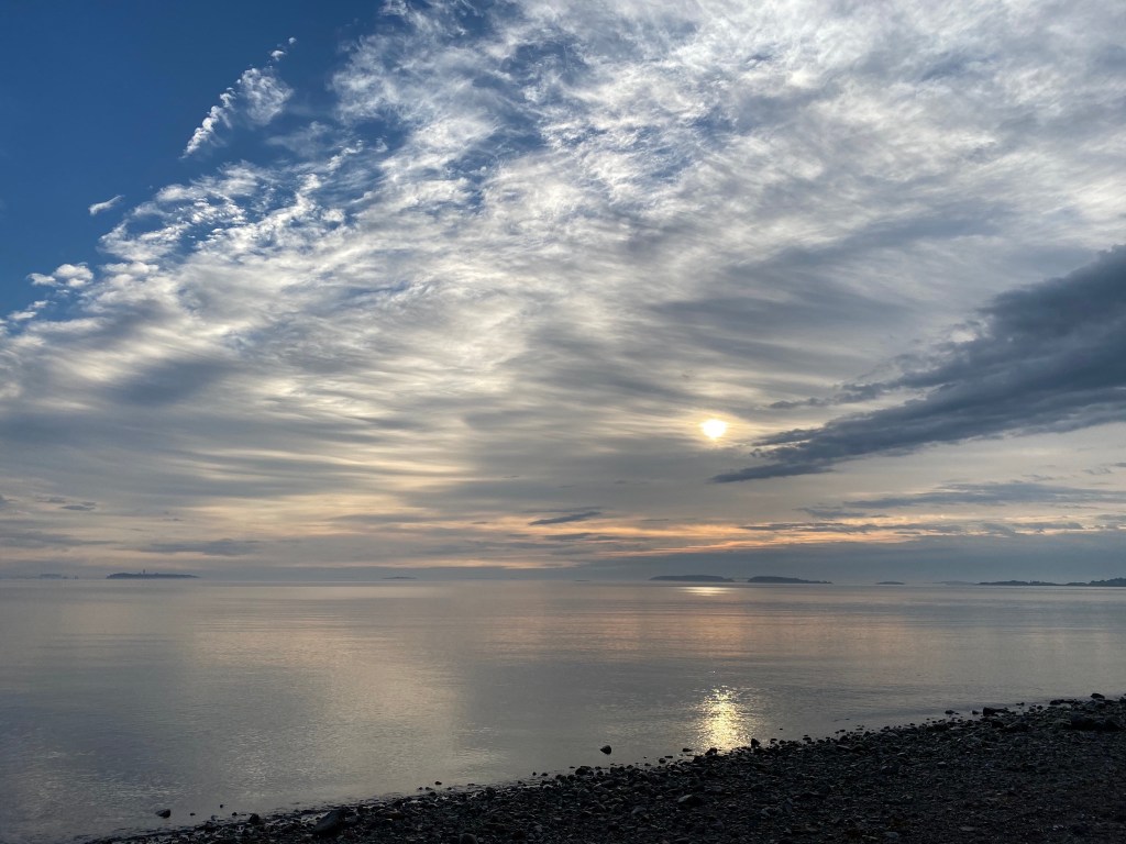 Photo is of a beach with a blue and cloudy sky with the sun and peach tones in the background.