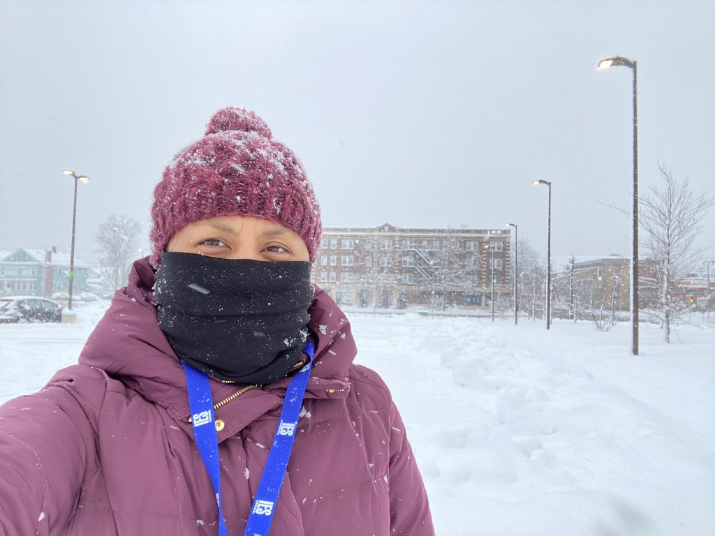Photo is of Lindsay taking a selfie and wearing a purple beanie, black gaiter that is covering her face from the nose and down, purple down coat, and blue lanyard with white text. It is snowing, and there is lots of snow covering the parking lot in the background. There are also a few buildings in the background.