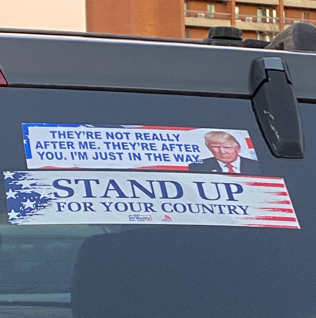 Photo is of two bumper stickers on a black car. The top one has an image of Donald Trump and says, "They're not really after me. They're after you. I'm just in the way." The bottom one says, "Stand up for your country."