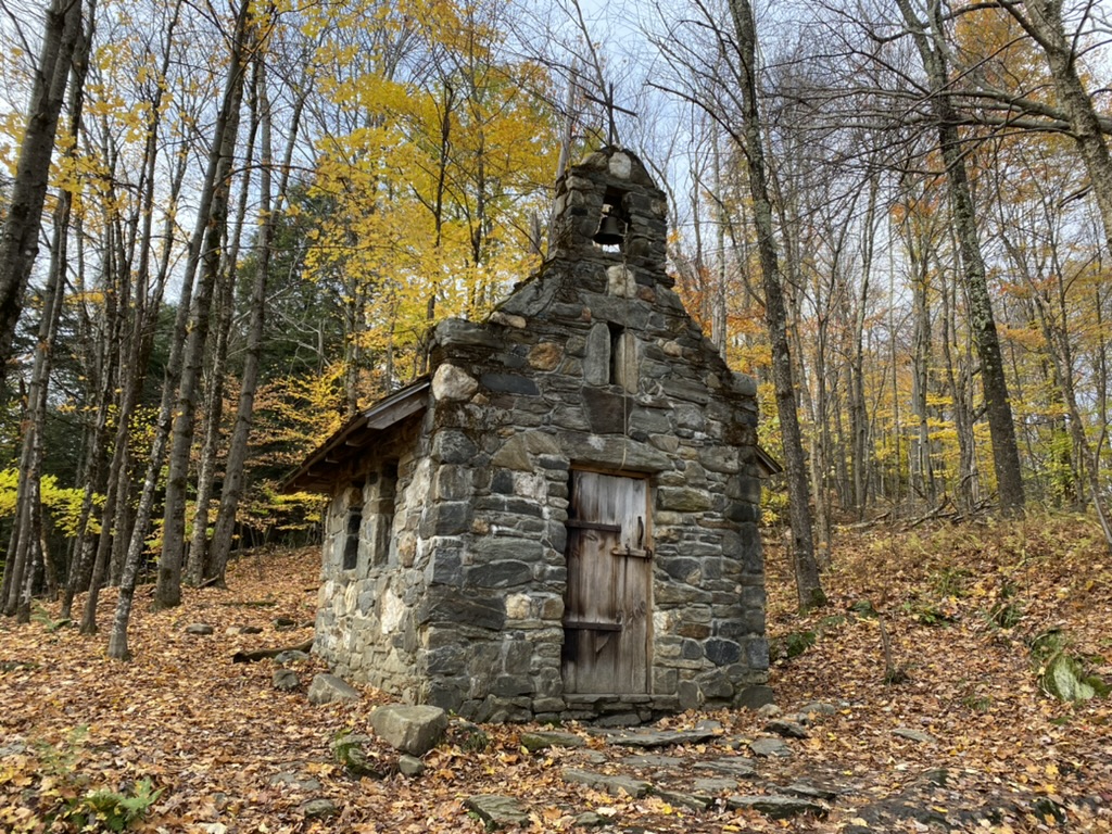 Photo is of a small stone chapel surrounded by trees mostly without leaves and one tree with yellow leaves. It is autumn. There are orange leaves covering the ground.