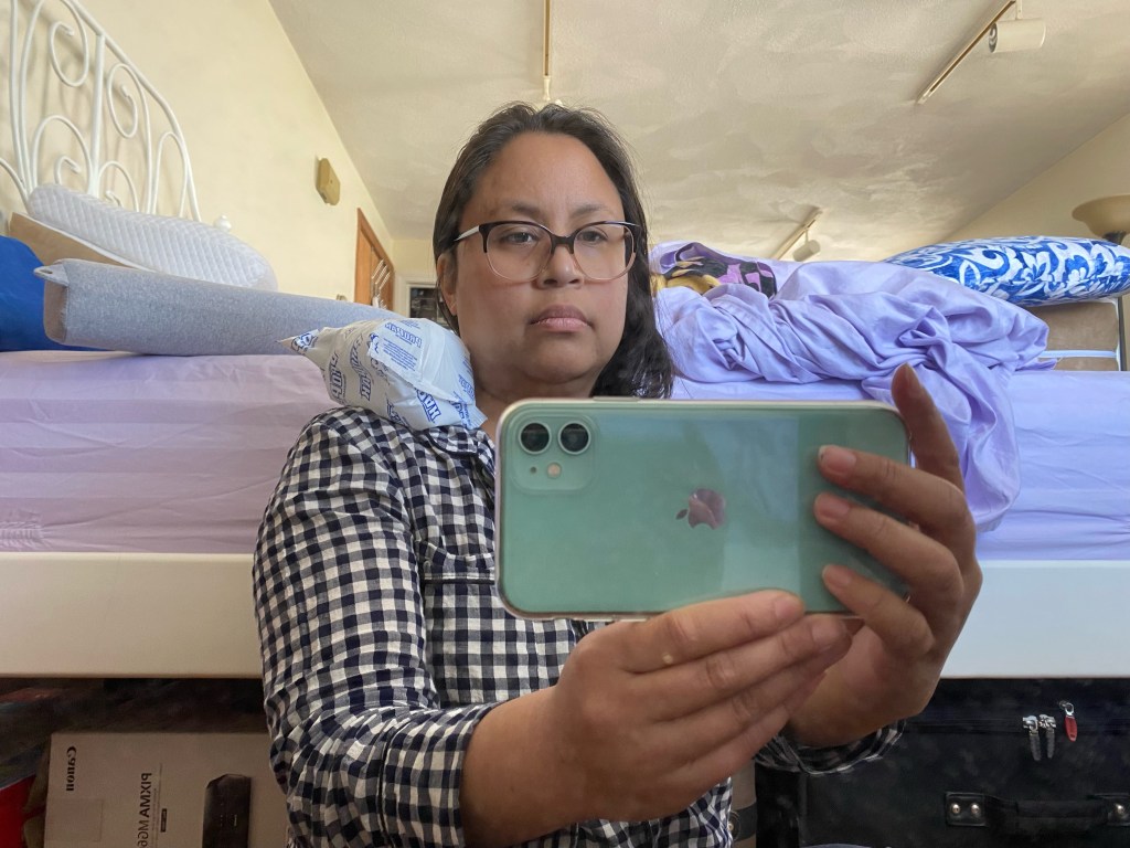 Photo is of a Brown woman taking a selfie of herself. She has an ice pack on her shoulder. She is wearing glasses and a pajama top with a navy blue and white plaid pattern.