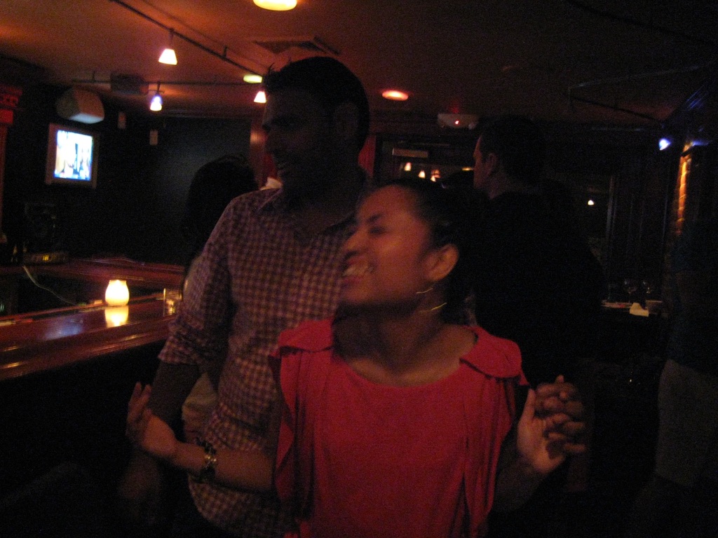 Photo of a Brown woman dancing and smiling in a blurry photo. There is a Brown man, whose face blends into the darkness, behind her.